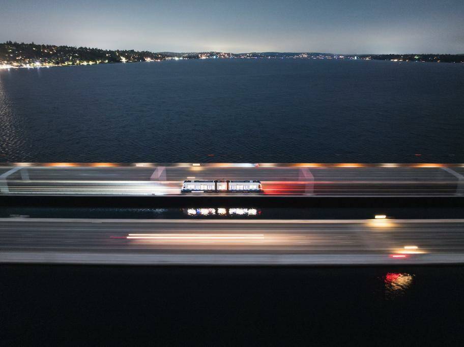 A one-car Sound Transit Link light rail train zooms across the Interstate 90 bridge at night, with car headlights blurring by. Photo from Sound Transit’s Platform blog