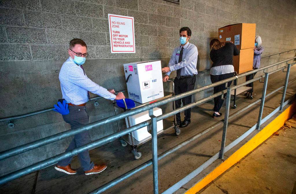 University of Washington Medical Center Montlake campus pharmacy administration resident Derek Pohlmeyer (left) and UWMC pharmacy director Michael Alwan transport a box containing Pfizer-BioNTech COVID-19 vaccines toward a waiting vehicle headed to the UW Medical Centers other hospital campuses on Monday in Seattle. (Mike Siegel/The Seattle Times via media pool)